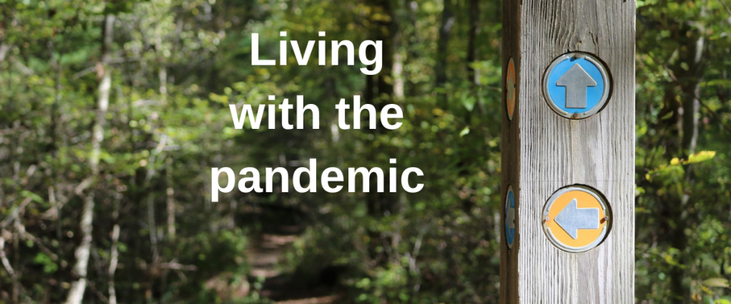 Image of a post which has signs pointing in the direction of various paths within a woodland area. The text says "Living with the pandemic".