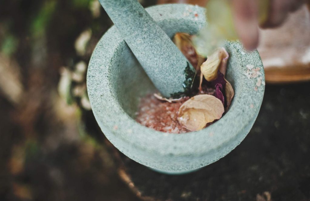 A stone pestle and mortar with some pink salts and beige and purple leaves.