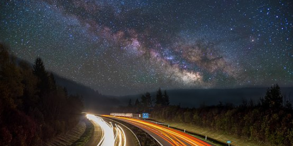 Overlooking a busy road with lines of light from the moving traffic with a starry night sky and trees lining the road.