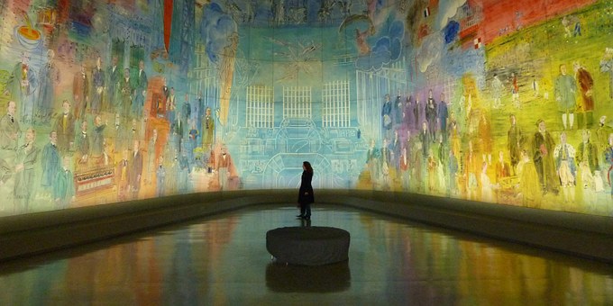 A woman looking at an art exhibition that covers all the walls and depicts historical buildings and people.
