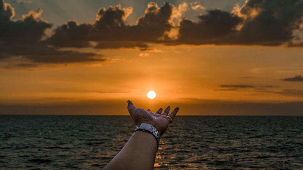An orange sunset above the sea where the sun is low on the horizon and an arm reaches out below the sun as if it were holding the sun on the palm of its hand.