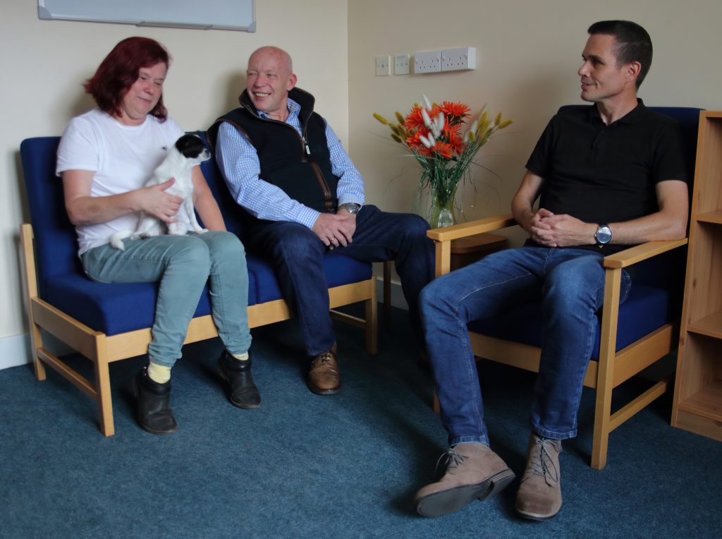 Woman with dog on her knee sitting talking with two men, all smiling.