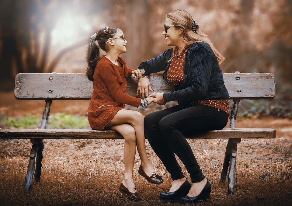 A woman and a child sat together on a park bench in autumn smiling at each other and holding hands.
