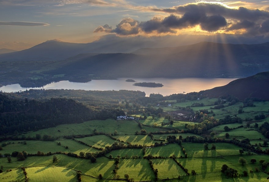 the sun shining through the clouds over a lake and green fields